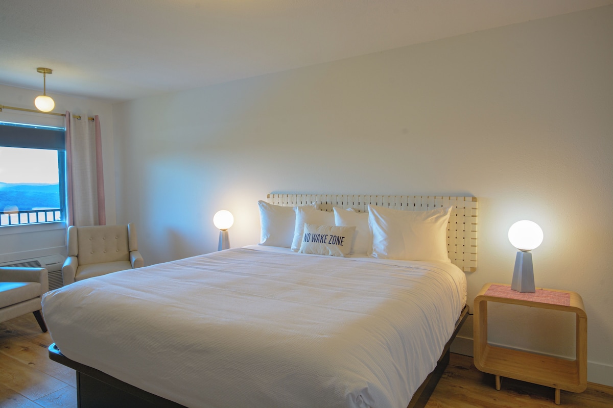 A hotel room with a large bed, two bedside lamps, a window with curtains, a chair, and a small wooden side table.