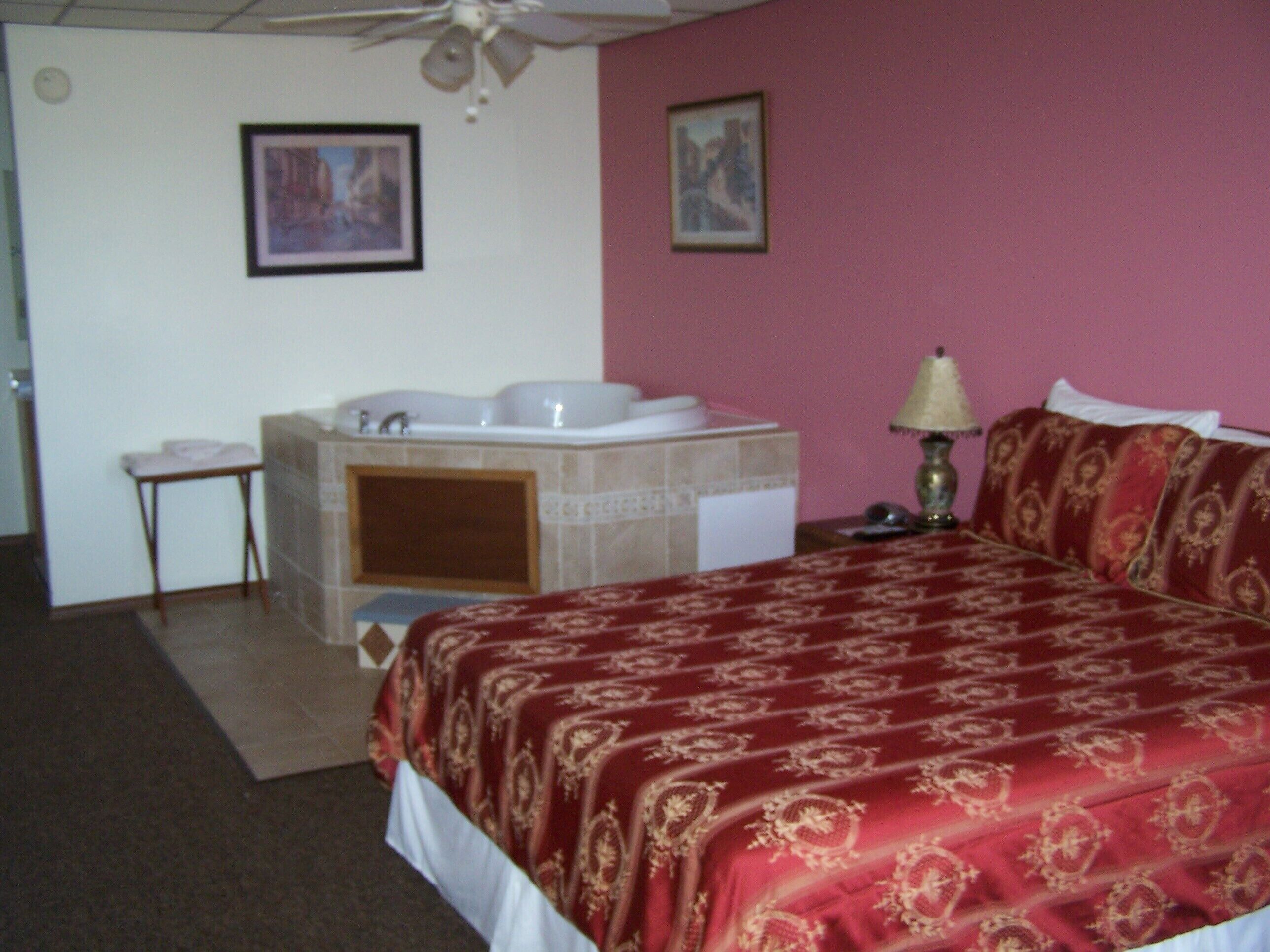 Quarto de hotel com cama, banheiro com banheira, uma mesinha e duas imagens emolduradas na parede.