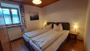 A double bed with two pillows, a nightstand with a lamp, a window with curtains, and a radiator. - Vacation apartment no. 1 (Neuötting)
