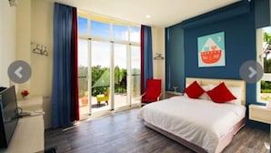 A hotel room with a large bed, a desk with a computer, a red chair, and a view of the outdoors through a sliding glass door. - Full Moon Resort (Hengchun)
