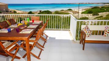 Un balcón con mesa de madera dispuesta para comer, un sofá con cojines estampados y vistas a la playa y al océano.