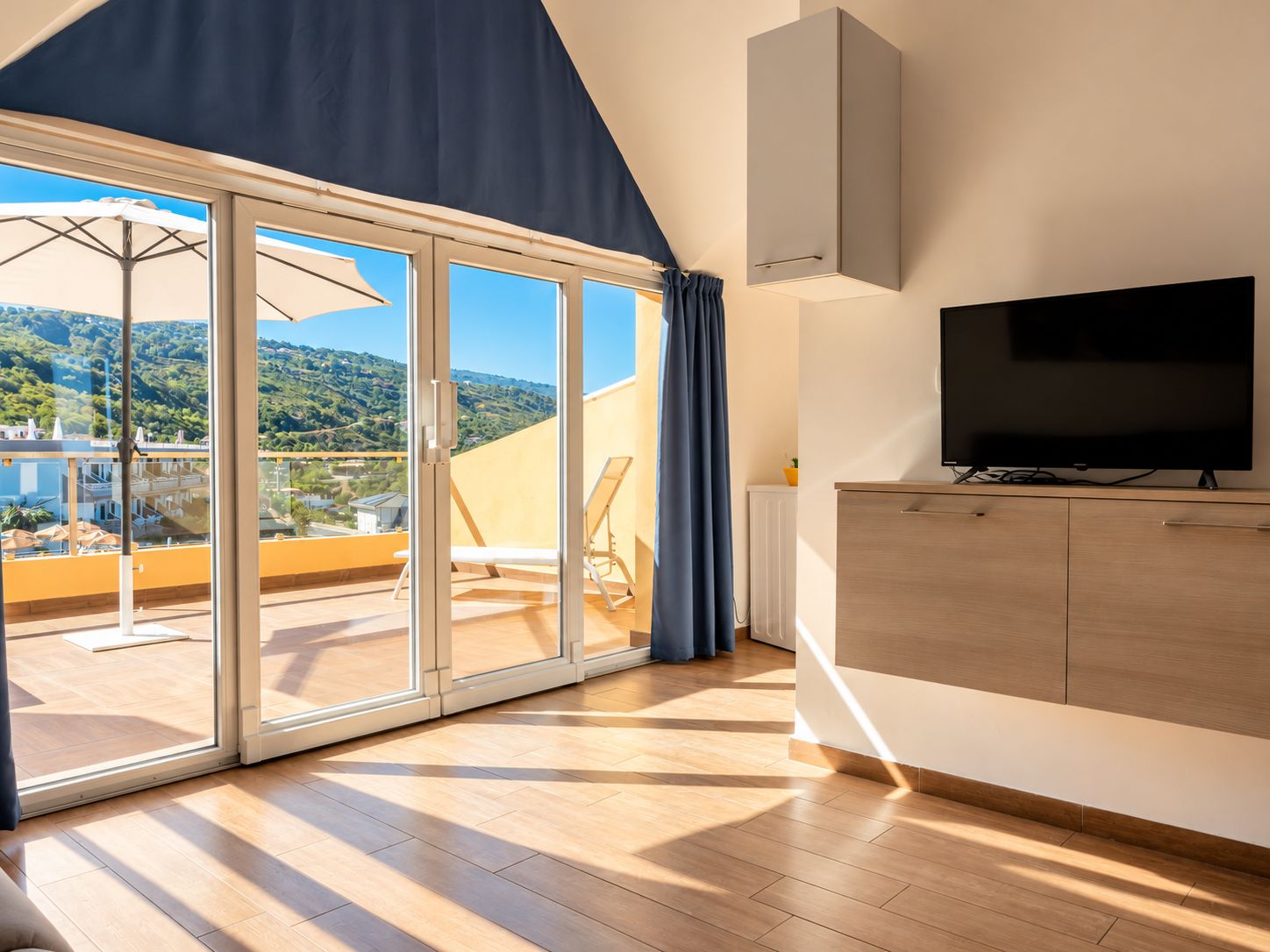 A modern room with a large sliding glass door leading to a balcony, a flat-screen TV mounted on the wall, and a wooden floor.