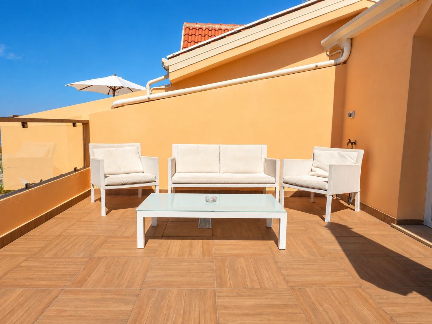 Una terraza en la azotea con un conjunto de sofá blanco, una mesa de centro de vidrio y piso de baldosas.