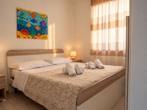 Ein Hotelzimmer mit einem Bett, einem Nachttisch, einer Lampe, einem Fenster mit Vorhängen und einem farbenfrohen Bild an der Wand.