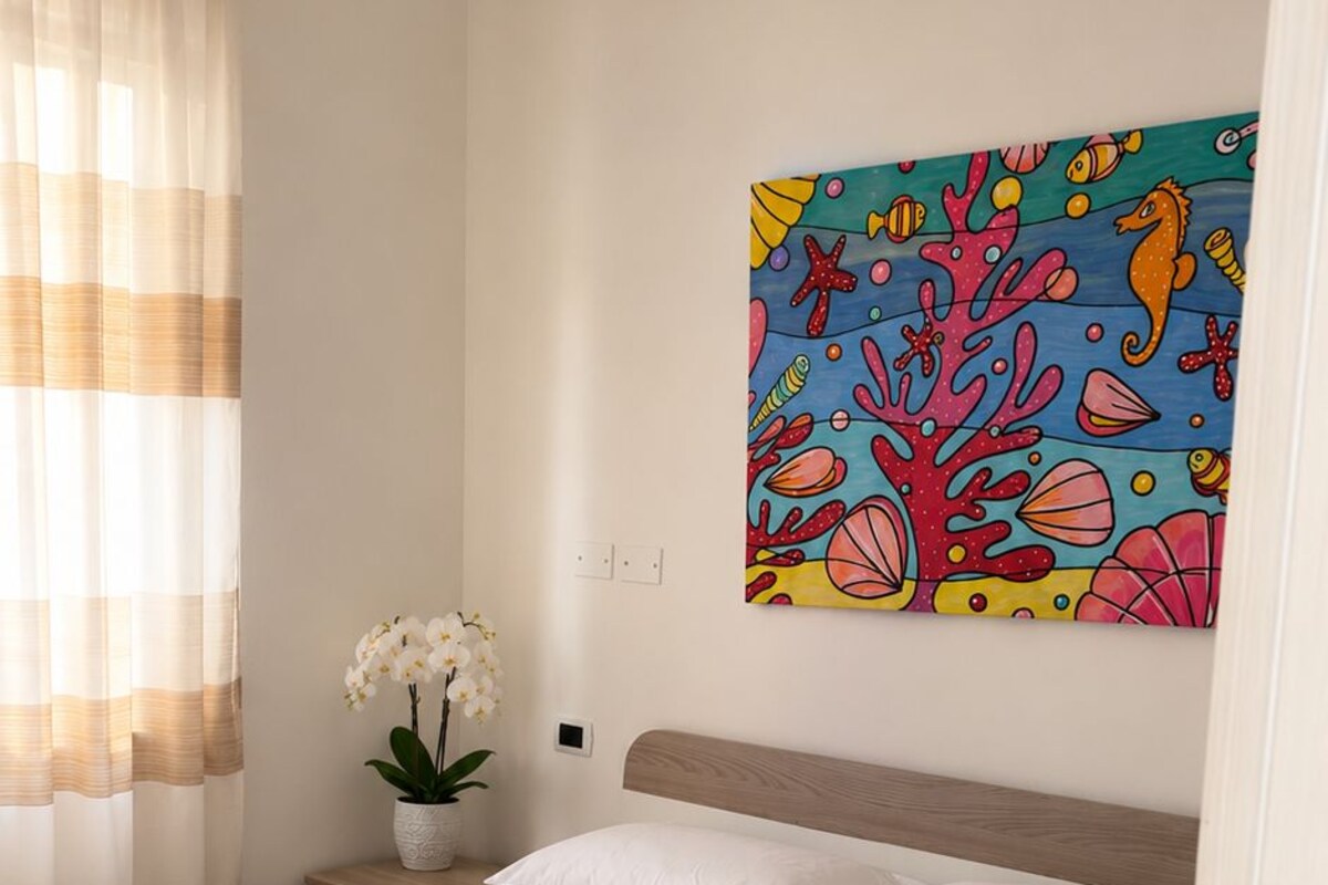 Un dormitorio con cama, ropa de cama blanca, colcha con estampado floral, mesita de noche con un jarrón de flores blancas y un cuadro colorido en la pared.
