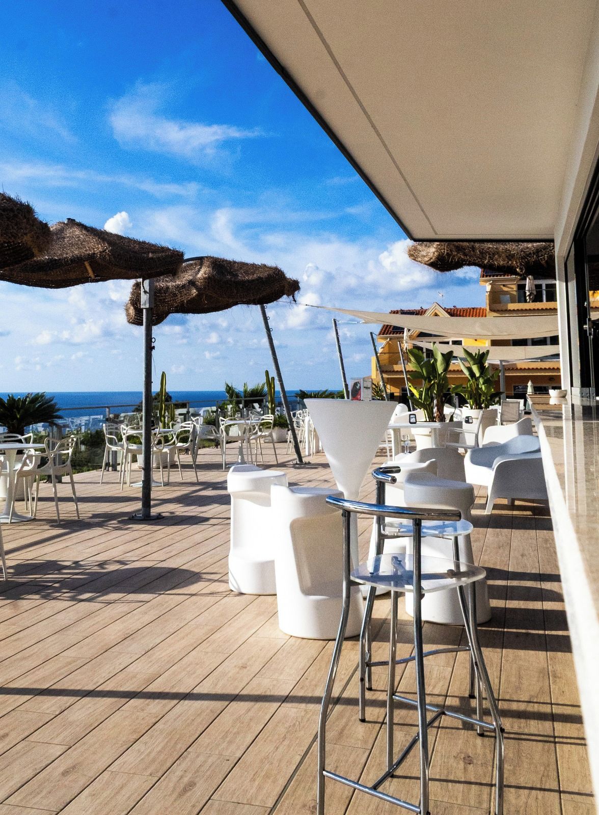Ein sonnendurchfluteter Außenbereich mit weißen Stühlen und Tischen, einer Holzterrasse und Blick auf das Meer.