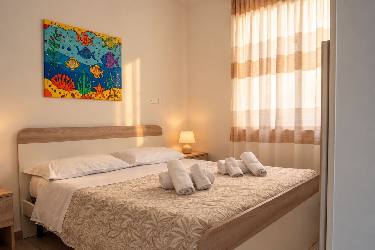 Habitación de hotel con cama, mesita de noche, lámpara, ventana con cortinas y un cuadro de colores en la pared.