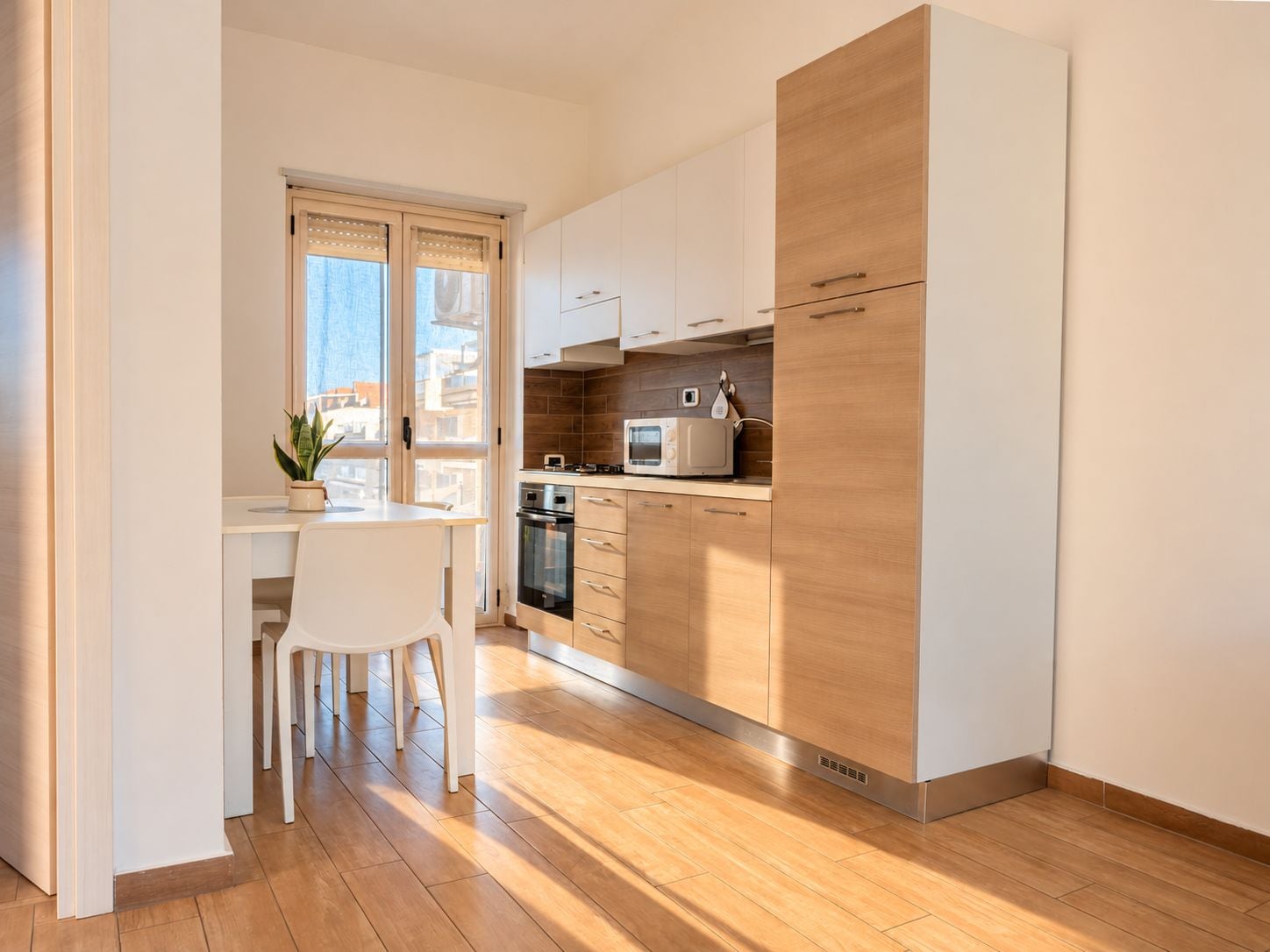 A modern kitchen with white cabinets, a microwave, and a stove. There is a dining area with a white chair and a plant.
