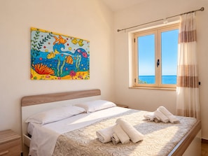A bedroom with a bed, a painting of fish, a window with a view of the sea, and towels on the bed.