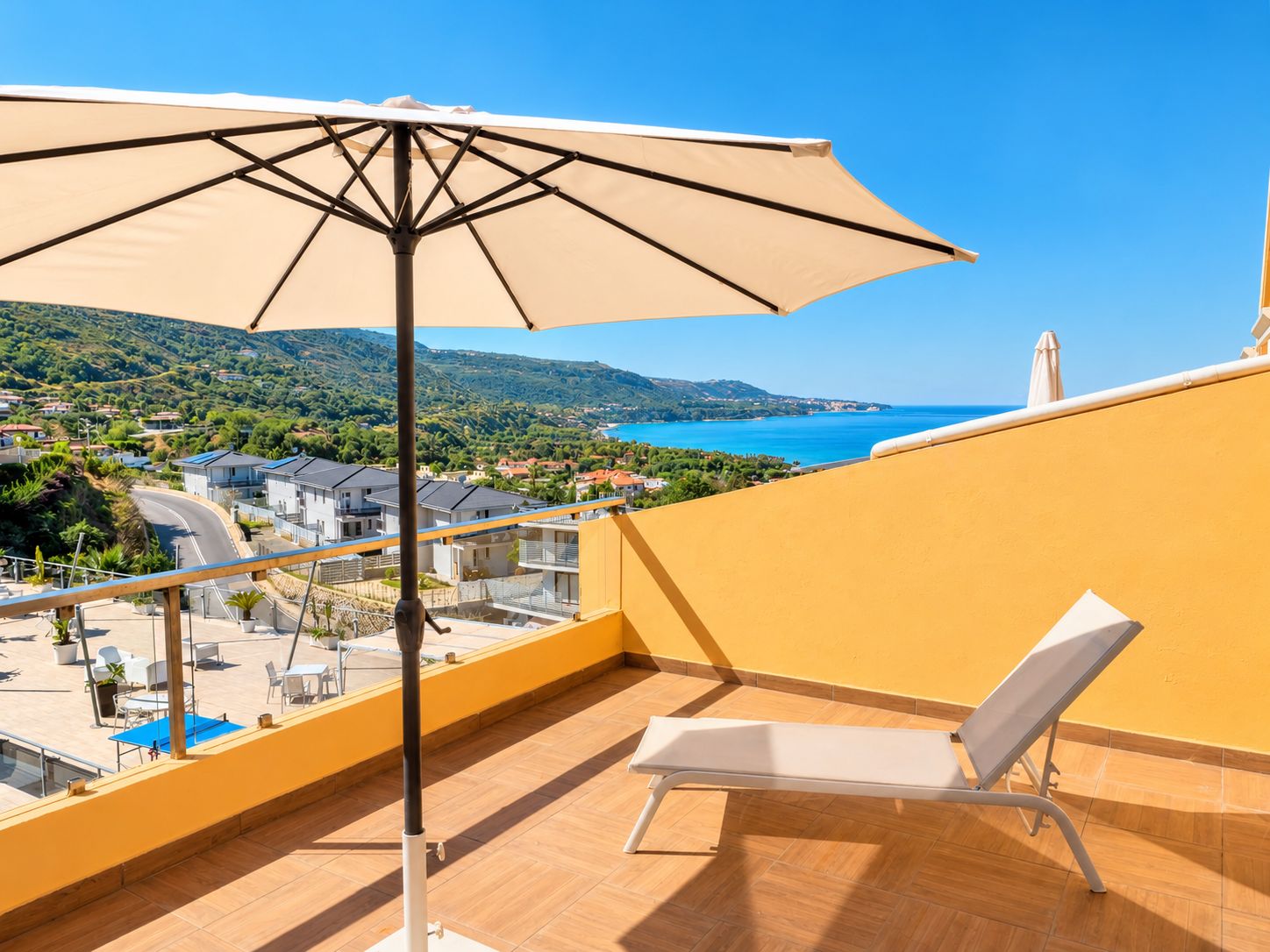 Un balcone con una poltrona bianca e un ombrellone beige, con vista su una cittadina costiera e sul mare.