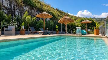 A clear swimming pool with lounge chairs and umbrellas, surrounded by greenery and a cliff.