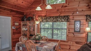 Interior acolhedor de uma cabana com uma mesa de jantar de madeira, cadeiras, uma estante de livros e uma janela com persianas.