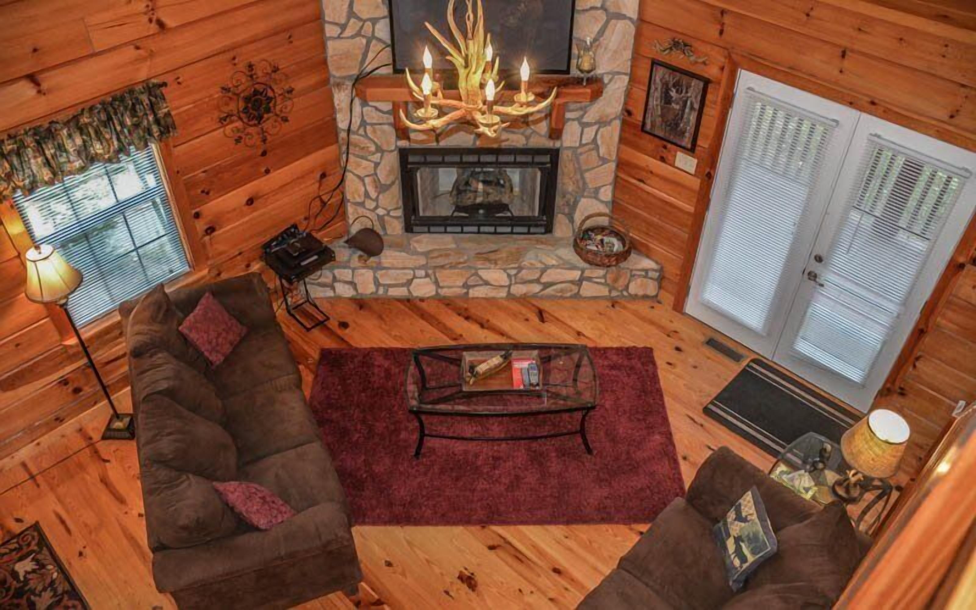 A cozy cabin interior with a stone fireplace, wooden walls, a sofa, and a large window with blinds.