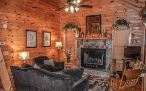 A cozy cabin interior with a fireplace, two armchairs, a television, and a ceiling fan.