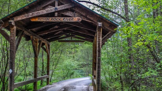 Een houten brug in rustieke stijl met een bord waarop 'Indian Rock River' staat, leidend naar een bebost gebied.