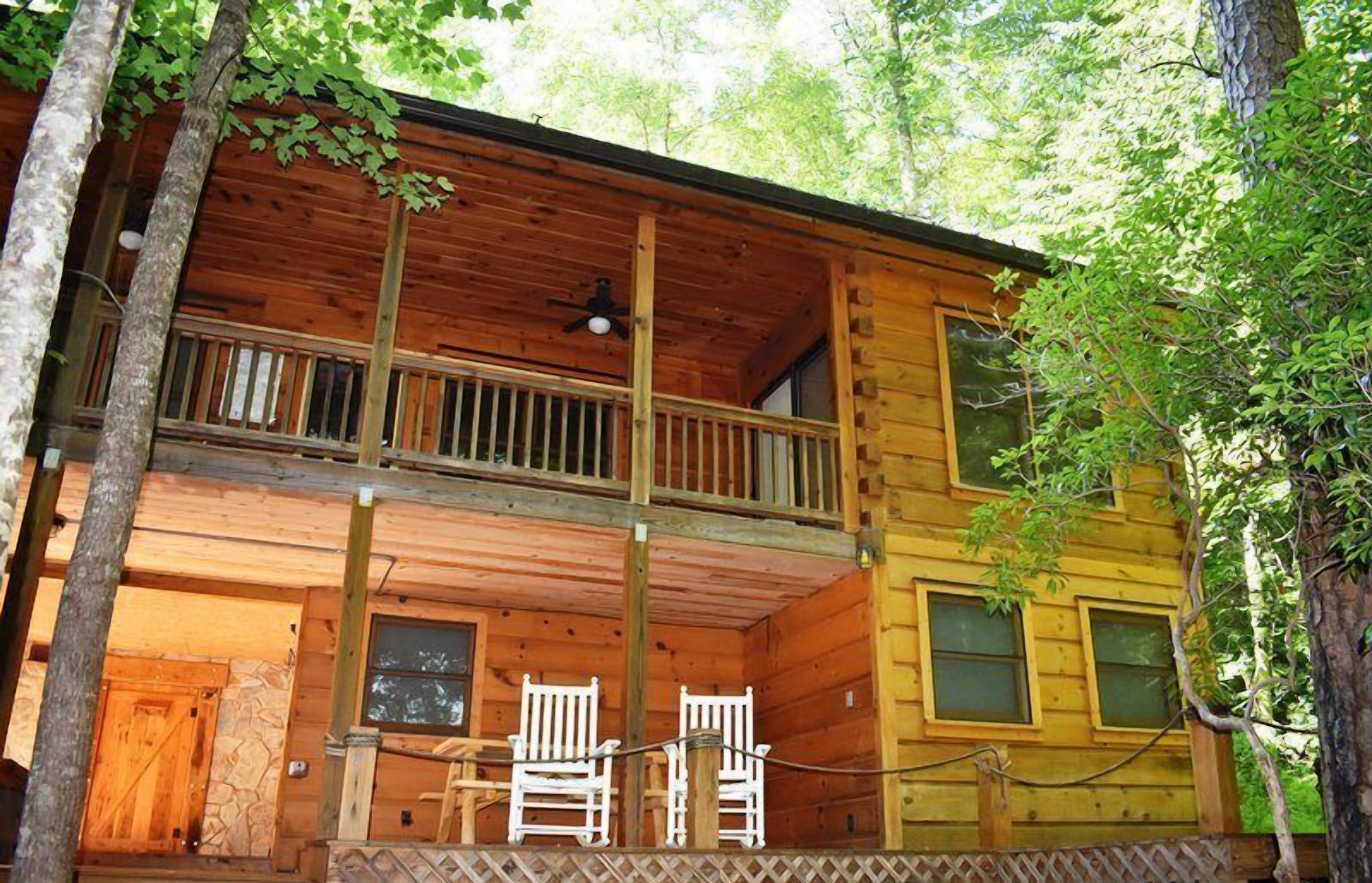 A two-story log cabin with a balcony, wooden railings, and a small porch area with chairs.