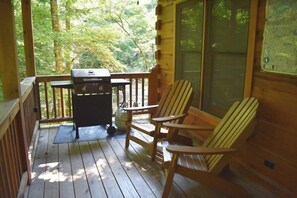 A wooden deck with a grill and Adirondack chairs, overlooking a forested area.