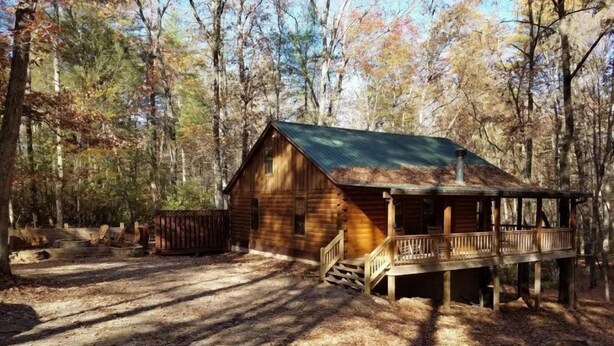 A rustic cabin with a porch and a covered deck, surrounded by a forested area.