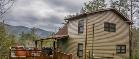 A two-story cabin with a wooden deck and a small porch, surrounded by trees and mountains.