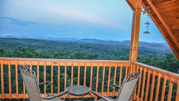 A wooden deck with two metal chairs and a small round table, overlooking a forested landscape with distant mountains. - Tree Tops (Mineral Bluff)