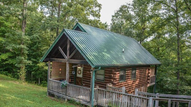 A rustic log cabin with a green metal roof, a porch with a railing, and a wooden deck. - Where The Maps End (McCaysville)