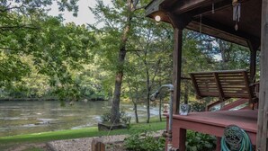 Una cabaña rústica con terraza de madera que da a un río, rodeada de un bosque denso.