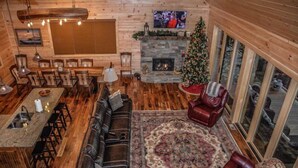 Een blokhutinterieur met een stenen open haard, een kerstboom, een grote televisie en een gezellige zithoek met vloerkleed en leren fauteuils.