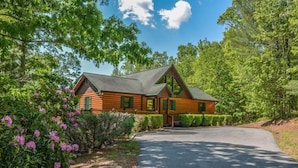 A log cabin with a sloped roof, surrounded by lush greenery and flowering bushes. - Bella Vista (Blue Ridge)