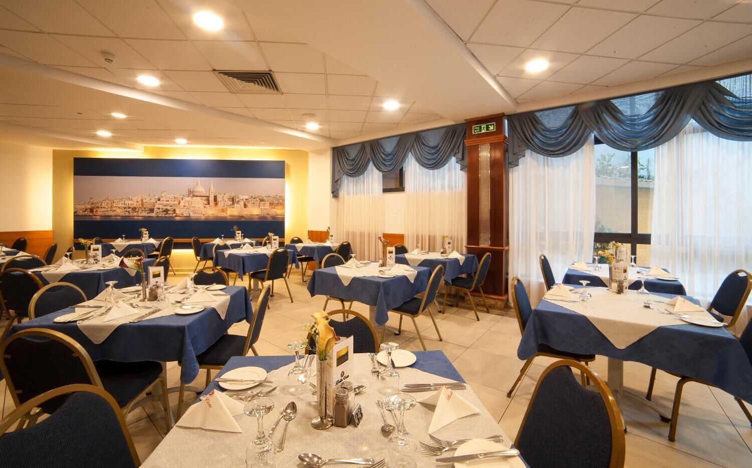 A dining area with tables set for service, a cityscape mural on the wall, and large windows with curtains.