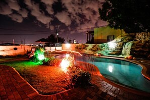 A swimming pool with a waterfall feature, surrounded by a brick pathway and illuminated plants. - Grand View (Morganton)