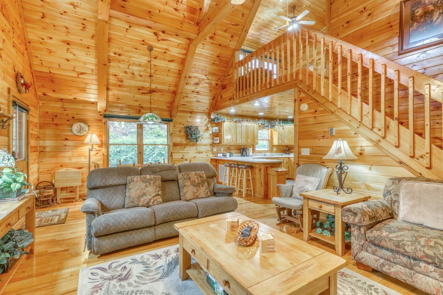 A cozy cabin interior with a wooden staircase, a living room with a sofa and chairs, a kitchen area, and a dining table.