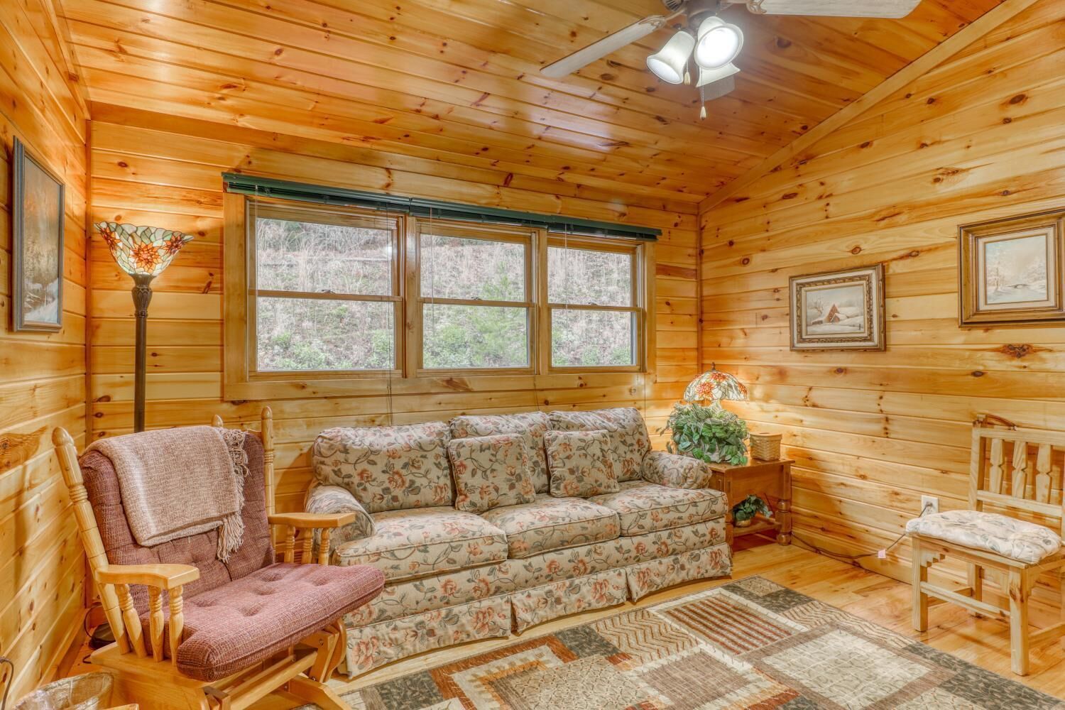 A cozy cabin interior with a floral sofa, wooden rocking chair, and a large window with a view of the outdoors.