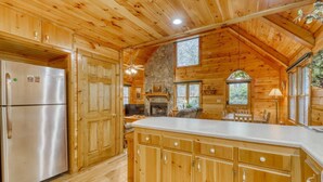 A wooden cabin interior with a kitchen featuring a stone fireplace, wooden cabinets, and a large island. - Lucilles Creekside Hideaway (Blue Ridge)