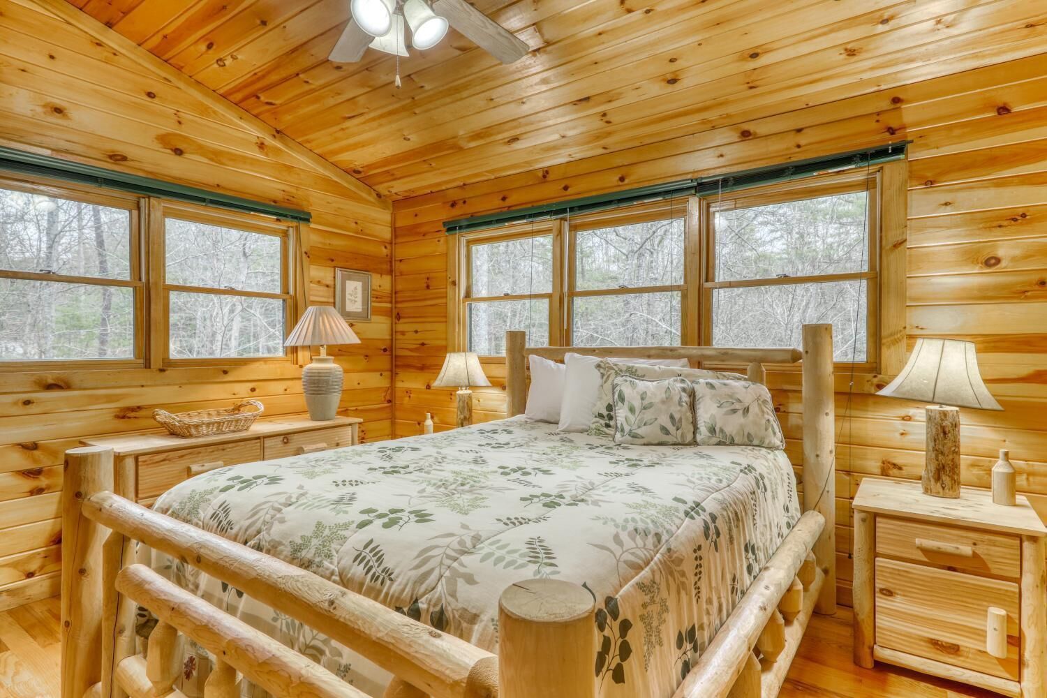 Une chambre à coucher dans une cabane en bois, avec un lit, deux tables de chevet, des lampes, une fenêtre et un ventilateur de plafond.