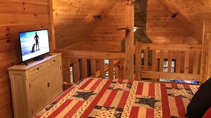 A wooden cabin room with a television on a cabinet, a quilt on the bed, and a stone fireplace. - Harleys River Retreat (Ellijay)