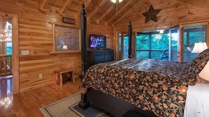 A wooden cabin bedroom with a bed, a ceiling fan, a star decoration, a television, and a window with a view of trees. - Panoramic Paradise (Morganton)