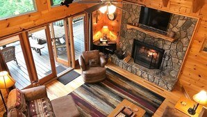 A cozy cabin interior with a stone fireplace, leather furniture, and a large TV screen. - Light`s Mountain Lake Overlook (Blue Ridge)