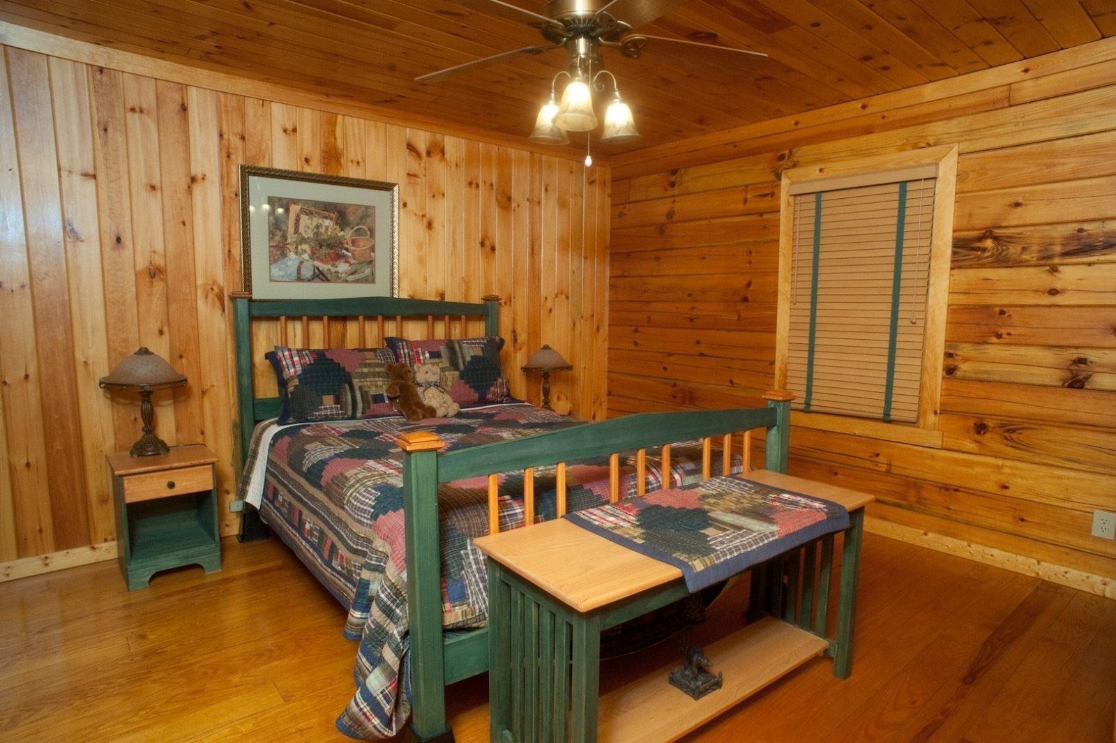 Une chambre à coucher située dans une cabane en bois, comprenant un lit, deux tables de chevet, un ventilateur de plafond et une fenêtre avec des stores.