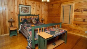 A wooden cabin bedroom with a bed, two bedside tables, a ceiling fan, and a window with blinds. - Three Bears Bluff (Cherry Log)
