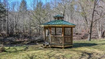 A wooden gazebo with a green roof, surrounded by a grassy area and trees.