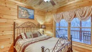 A wooden cabin bedroom with a bed, a ceiling fan, a painting, and a window with curtains. - The Bella Rose Retreat (Blue Ridge)
