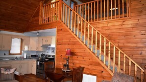 A cozy cabin interior with a wooden staircase, a kitchen with basic appliances, and a dining area with a table and chairs.