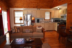 A cozy living room with a sofa, coffee table, and dining table. There is a kitchen area with cabinets and appliances.