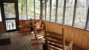 A wooden deck with rocking chairs and a bench, large windows with a view of trees, and a door with a tree design.