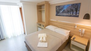 A hotel room with a bed, a bedside table with a phone, a wall-mounted light, a painting, and a wooden shelf unit. - Pousada Nascer do Sol (Arraial do Cabo)