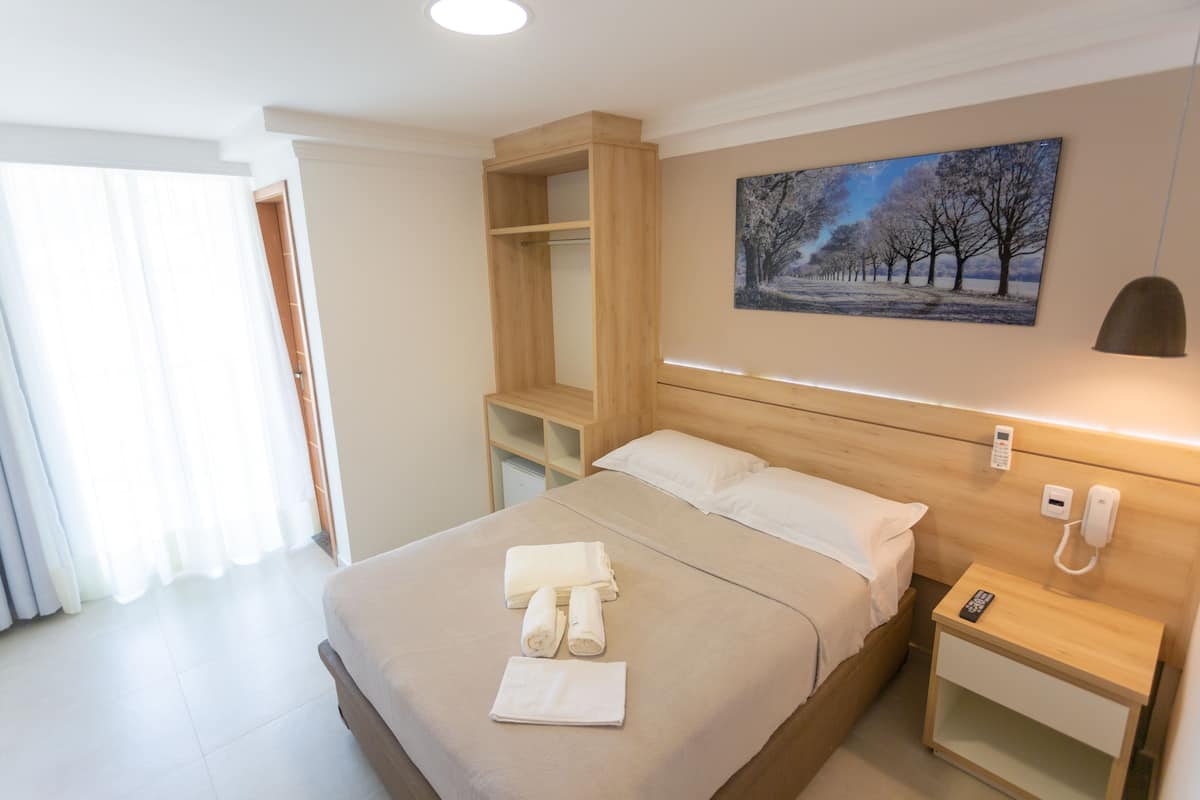 A hotel room with a bed, a bedside table with a phone, a wall-mounted light, a painting, and a wooden shelf unit.
