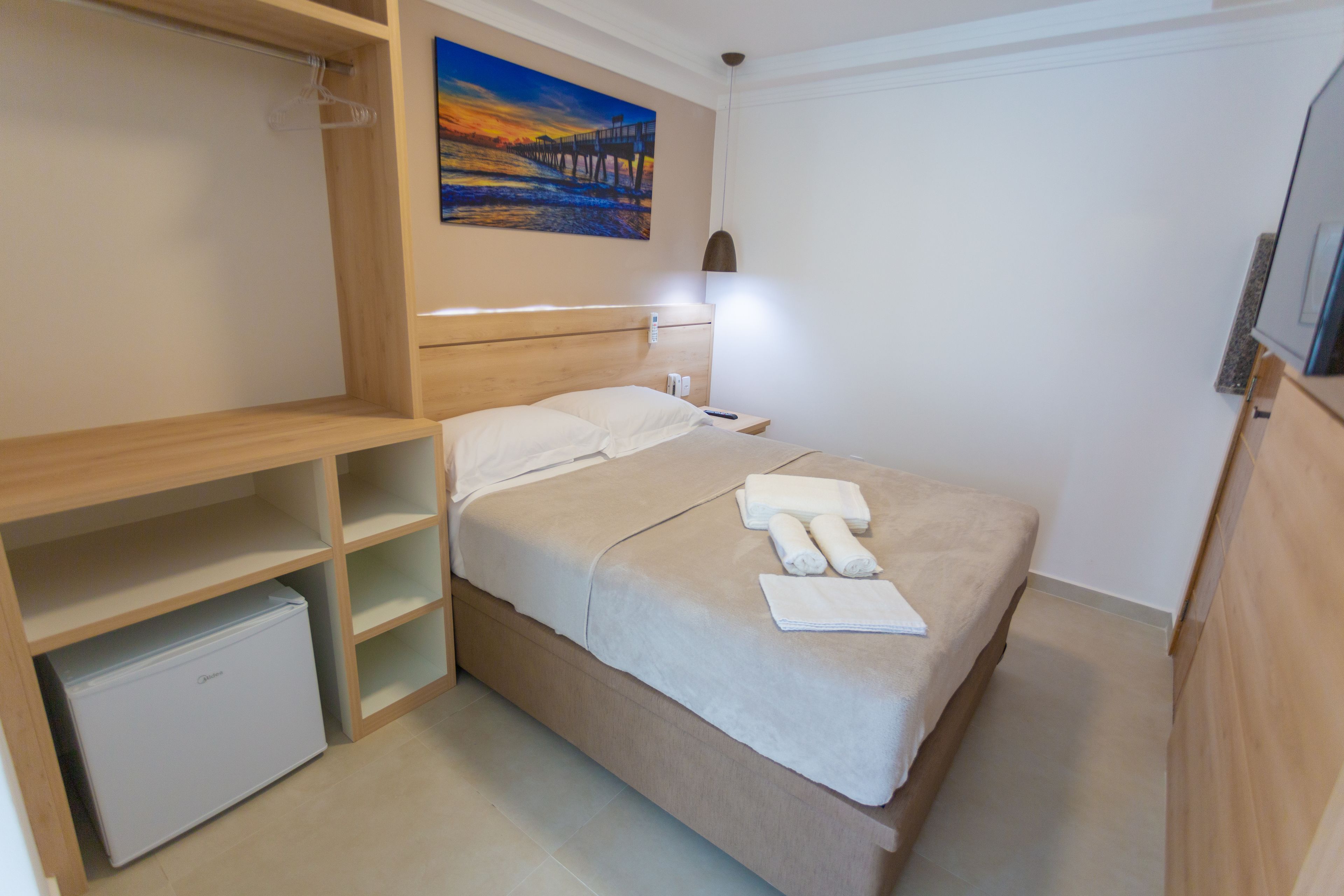 A neatly arranged bedroom with a bed, a small refrigerator, a painting on the wall, and a desk with a lamp.