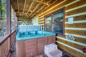 A wooden cabin with a hot tub on the porch, a window with a safety sign, and a stone wall in the background.