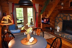 A cozy cabin interior with a dining table, chairs, a fireplace, a television, and a view of the outdoors. - Acorn Bungalow (Blue Ridge)