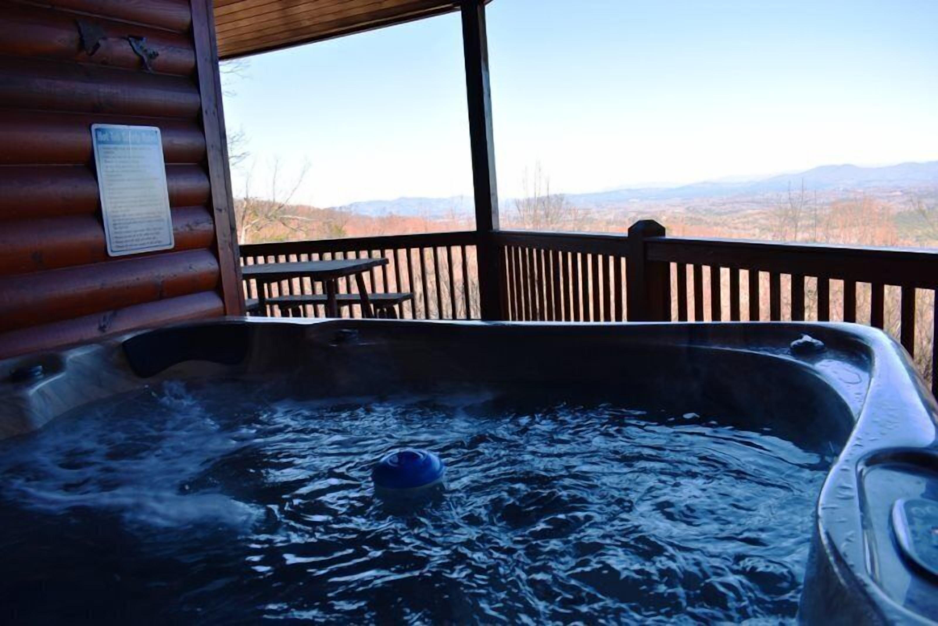 A hot tub with a wooden deck and a table, overlooking a mountainous landscape.